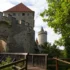 Kamenná hradní zeď s kulatou věží a červenou taškovou střechou, pohled z dřevěné lávky mezi zelenými stromy.