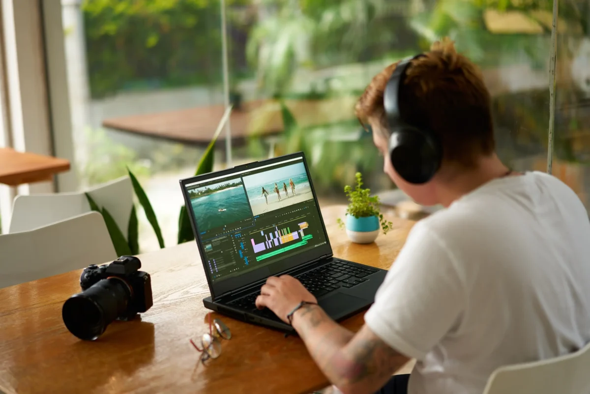 shutterstock_2454345013-změněno-na-3000x2004 | TVGURU.cz Osoba se sluchátky pracuje na projektu střihu videa na notebooku u dřevěného stolu s kamerou a brýlemi poblíž. Z okna za ní je vidět jasně zelená venkovní scenérie.