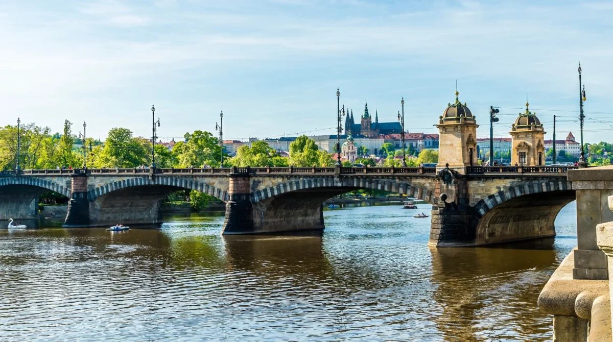 Pražský Karlův most přes řeku Vltavu s klenutými polemi, zdobenými lampami a vzdáleným panoramatem města.