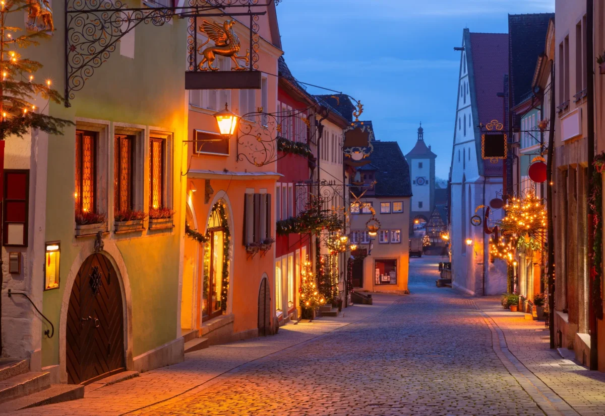 shutterstock_2695253443-změněno-na-3000x2067 | TVGURU.cz Dlážděná ulice v barevném evropském městě za soumraku, teplé lucerny a ozdobné železné cedule osvětlují scénu.