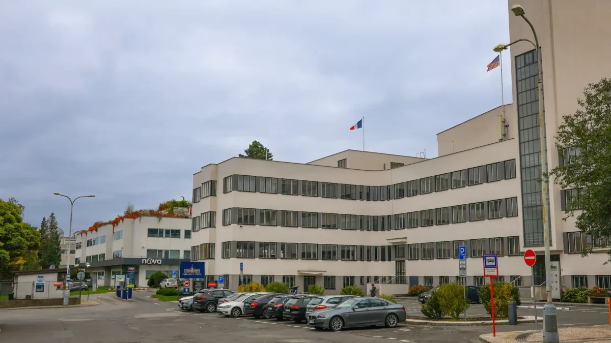 Modern white office building with many windows, two flagpoles (French and American) and a front parking lot under a cloudy sky.