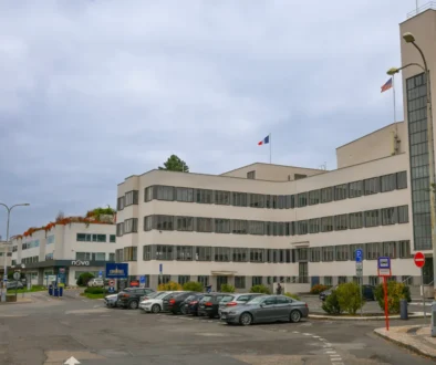 Modern white office building with many windows, two flagpoles (French and American) and a front parking lot under a cloudy sky.