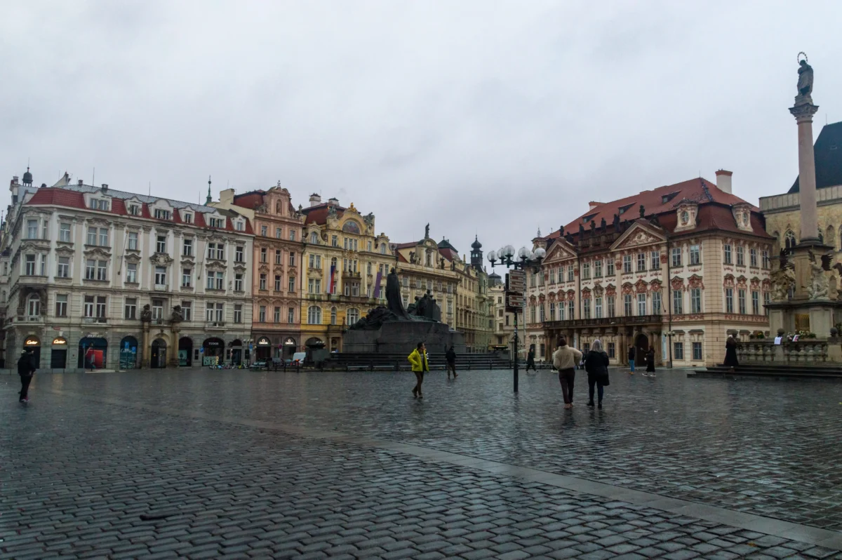 shutterstock_2713819945 1 | TVGURU.cz Historic square with pastel, ornate buildings along a cobblestone plaza; a statue in the foreground and a tall column statue on the right under a cloudy sky.