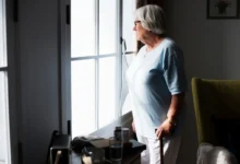 Seniorka stojí ve svém bytě a dívá se z velkého okna.