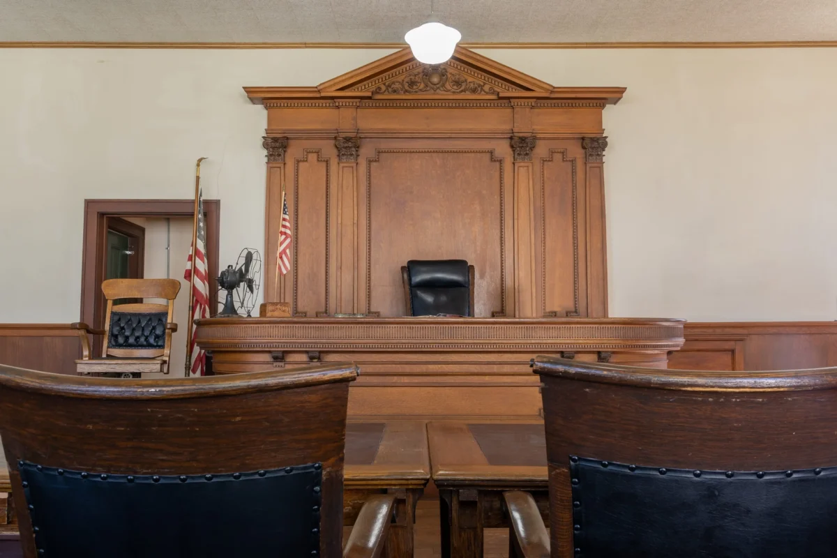 shutterstock_2298224579 1 | TVGURU.cz Wood-paneled judge's bench in a courtroom with flags and empty seating in front of it.