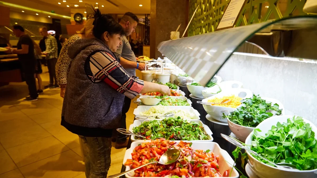 People serving themselves salad from a bright buffet line with bowls of greens and toppings at a restaurant or cafeteria.