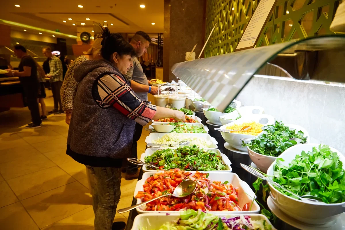 People serving themselves salad from a bright buffet line with bowls of greens and toppings at a restaurant or cafeteria.
