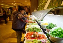 People serving themselves salad from a bright buffet line with bowls of greens and toppings at a restaurant or cafeteria.