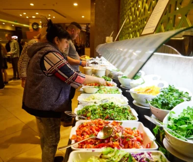 People serving themselves salad from a bright buffet line with bowls of greens and toppings at a restaurant or cafeteria.