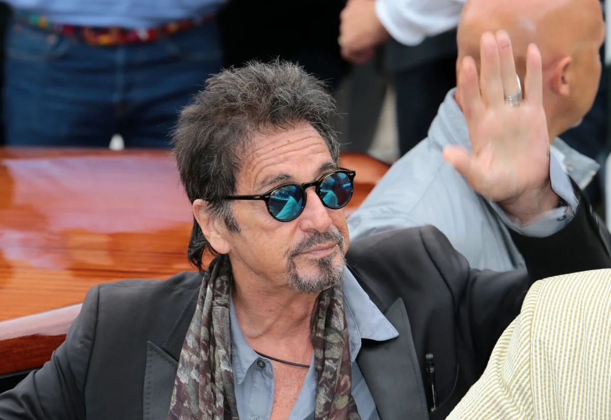 Man with gray facial hair wearing sunglasses and a scarf raises his left hand while seated at a wooden table or bench.