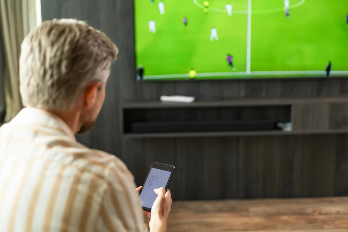 Změny pravděpodobně nastanou i u televizních programů. Zdroj: Shutterstock | TVGURU.cz Muž sedící u televize s telefonem v ruce