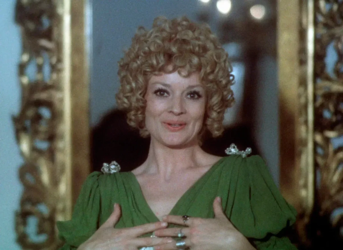 Woman with curly blond hair in a green dress, clasping her hands at chest and smiling, standing before a gilded frame.",
