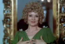 Woman with curly blond hair in a green dress, clasping her hands at chest and smiling, standing before a gilded frame.",