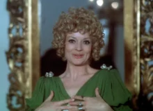 Woman with curly blond hair in a green dress, clasping her hands at chest and smiling, standing before a gilded frame.",
