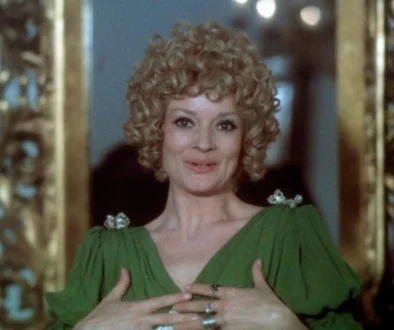Woman with curly blond hair in a green dress, clasping her hands at chest and smiling, standing before a gilded frame.",