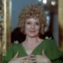 Woman with curly blond hair in a green dress, clasping her hands at chest and smiling, standing before a gilded frame.",