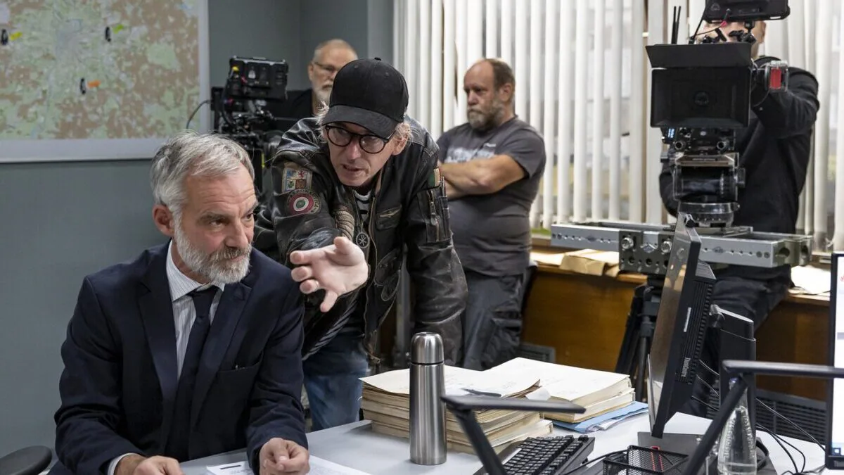 A suited man leans over a cluttered desk filled with papers as another man in a leather jacket points toward him, with cameras and a map in the background.