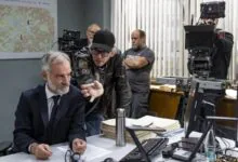 A suited man leans over a cluttered desk filled with papers as another man in a leather jacket points toward him, with cameras and a map in the background.