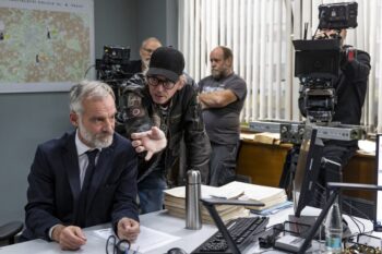 A suited man leans over a cluttered desk filled with papers as another man in a leather jacket points toward him, with cameras and a map in the background.