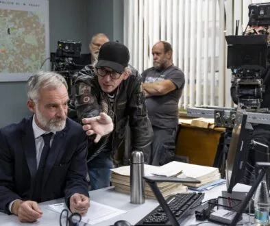 A suited man leans over a cluttered desk filled with papers as another man in a leather jacket points toward him, with cameras and a map in the background.