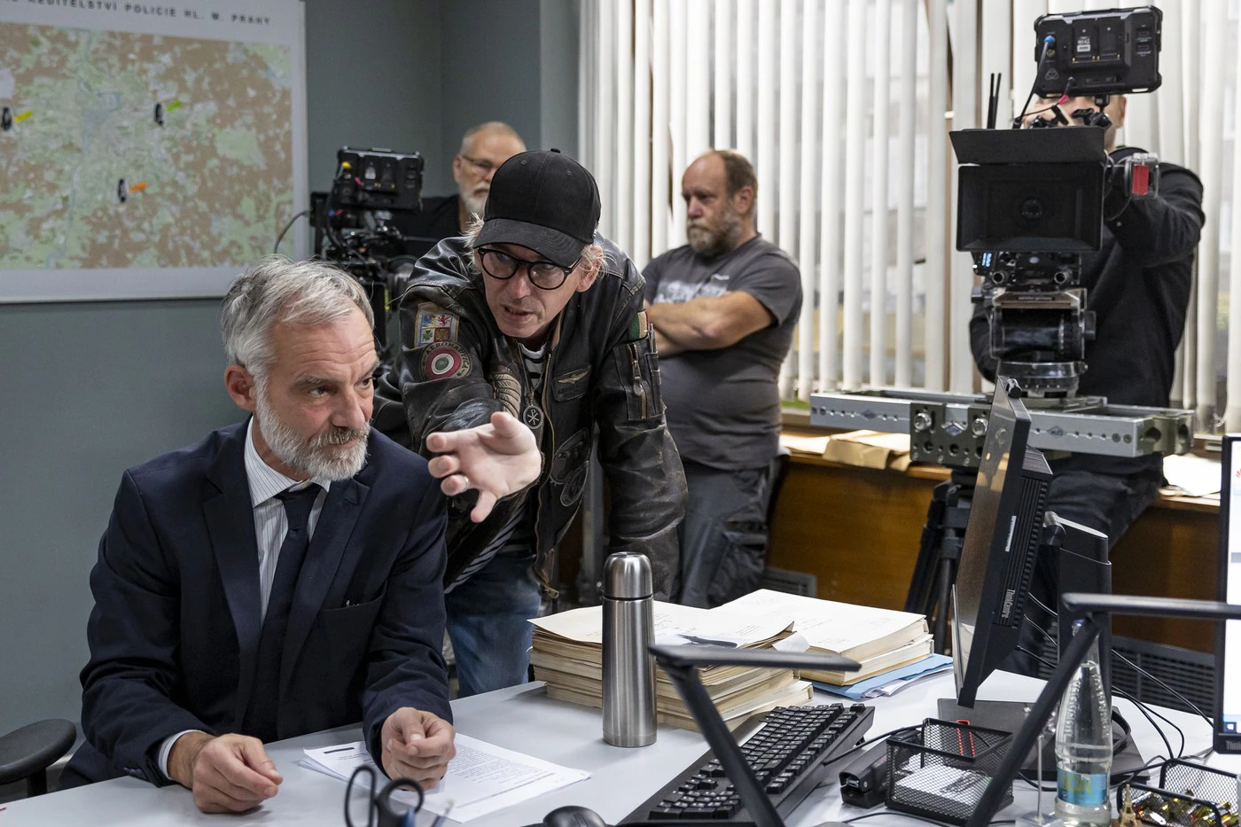 A suited man leans over a cluttered desk filled with papers as another man in a leather jacket points toward him, with cameras and a map in the background.