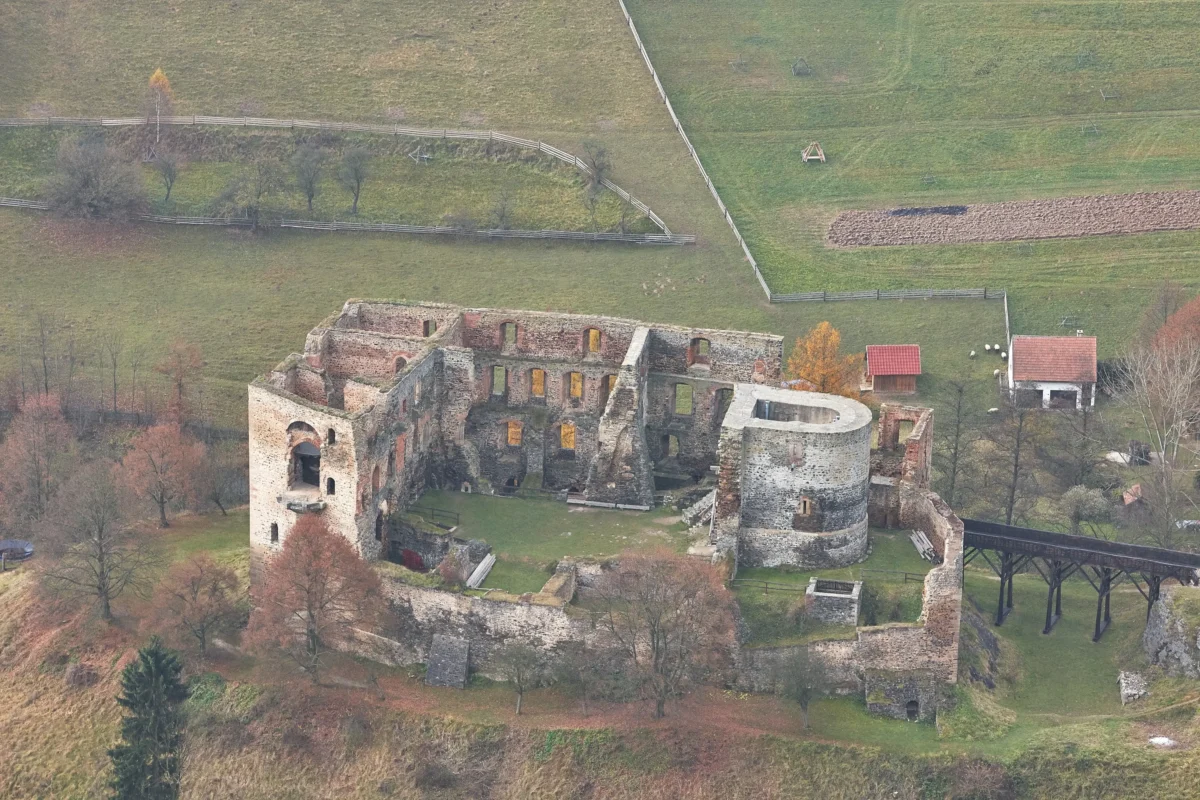 Letecký pohled na zříceninu středověkého kamenného hradu obklopenou zelenými poli a dřevěnými ploty