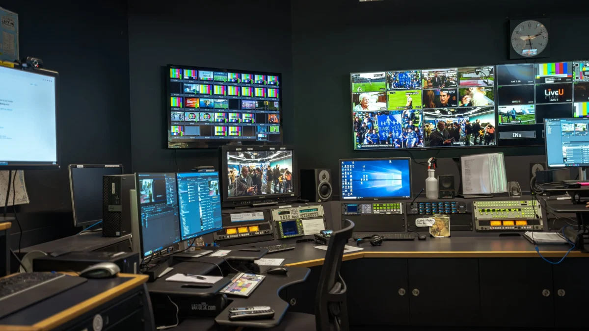 Broadcast control room with numerous screens displaying news feeds and control panels behind a desk setup.