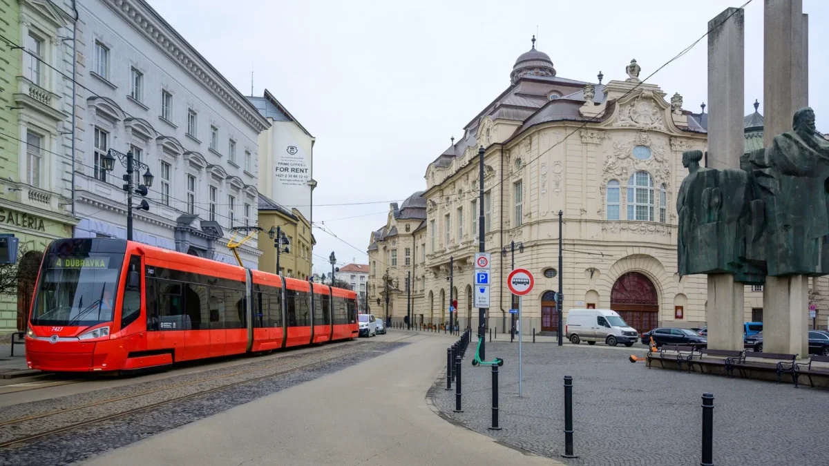 Red tram travels along a cobblestone street beside ornate historic buildings and a large statue on the right.