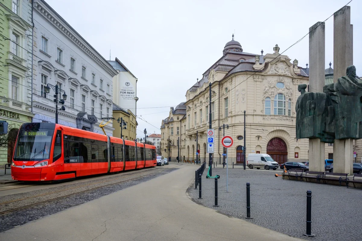 Red tram travels along a cobblestone street beside ornate historic buildings and a large statue on the right.