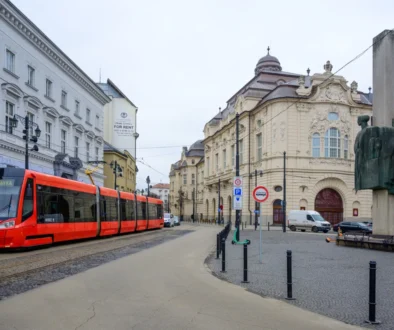 Red tram travels along a cobblestone street beside ornate historic buildings and a large statue on the right.