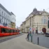 Red tram travels along a cobblestone street beside ornate historic buildings and a large statue on the right.