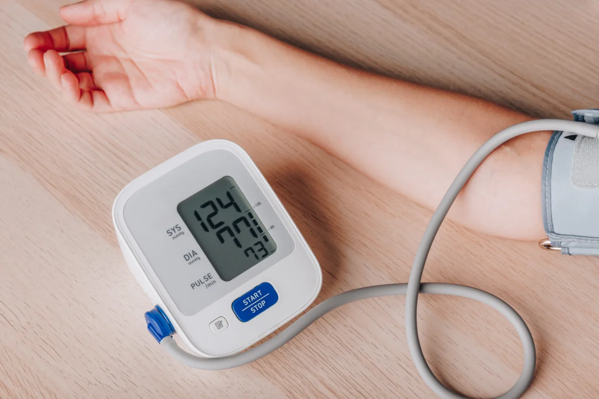 Arm with a cuff connected to a digital blood pressure monitor showing 124/70 mmHg and a blue Start/Stop button nearby, on a wooden surface