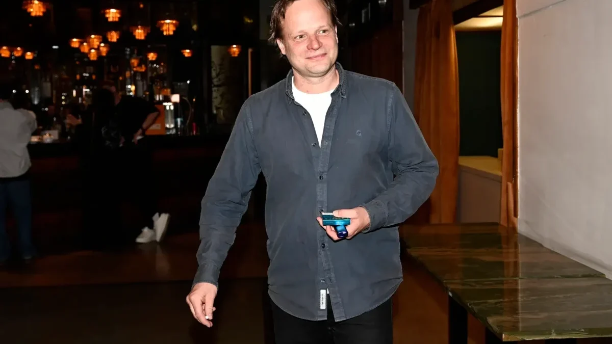 Smiling man in a dark blue shirt holding a small blue device in a dimly lit bar area.