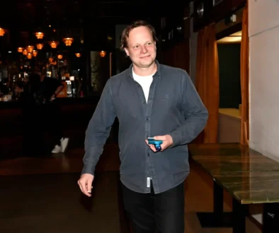 Smiling man in a dark blue shirt holding a small blue device in a dimly lit bar area.