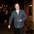Smiling man in a dark blue shirt holding a small blue device in a dimly lit bar area.