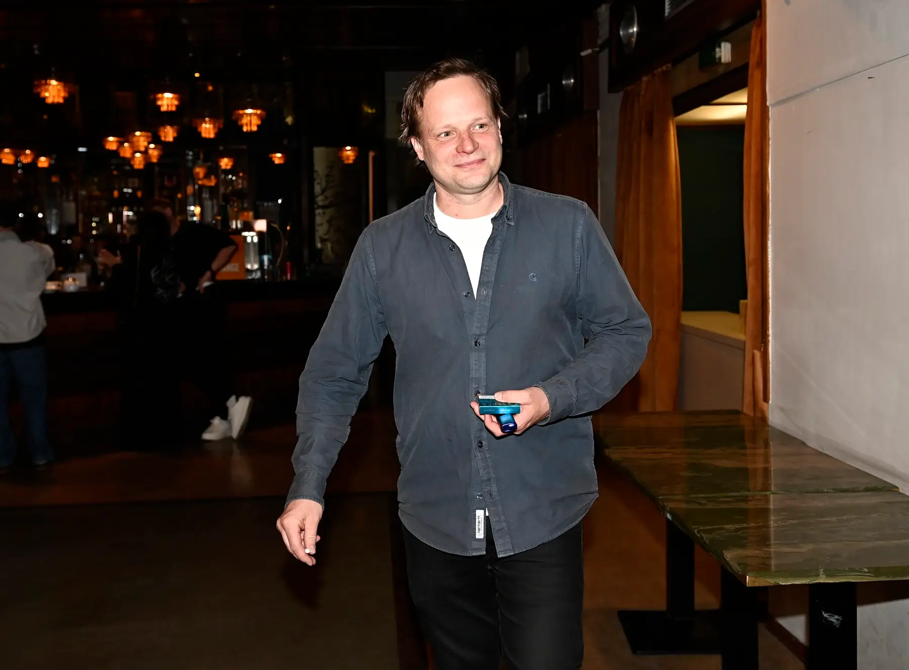 Smiling man in a dark blue shirt holding a small blue device in a dimly lit bar area.