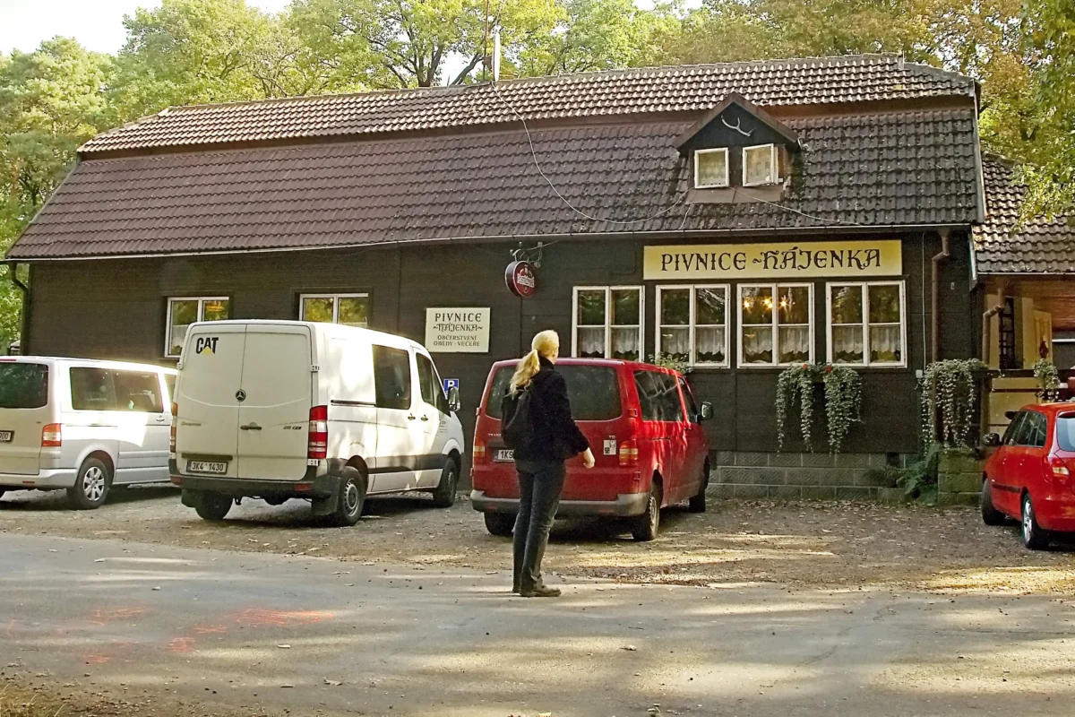 Tmavý dřevěný hostinec se žlutou cedulí s nápisem „Pivnice Kajenka“, před ním zaparkované bílé a červené dodávky a kolem procházející osoba.