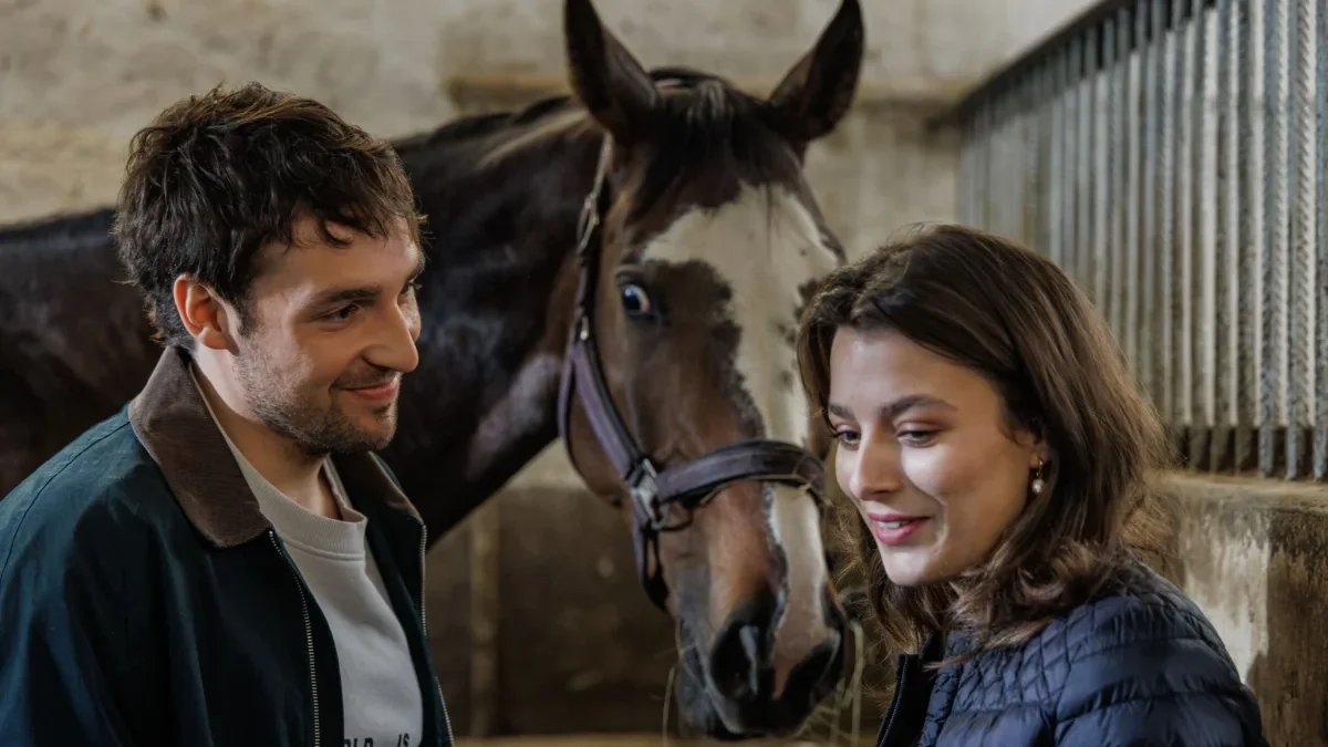 Man and woman smiling at a horse in a stable, with the horse between them in a barn setting.