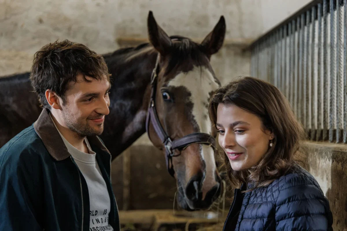 Man and woman smiling at a horse in a stable, with the horse between them in a barn setting.