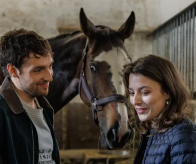 Man and woman smiling at a horse in a stable, with the horse between them in a barn setting.