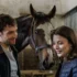 Man and woman smiling at a horse in a stable, with the horse between them in a barn setting.