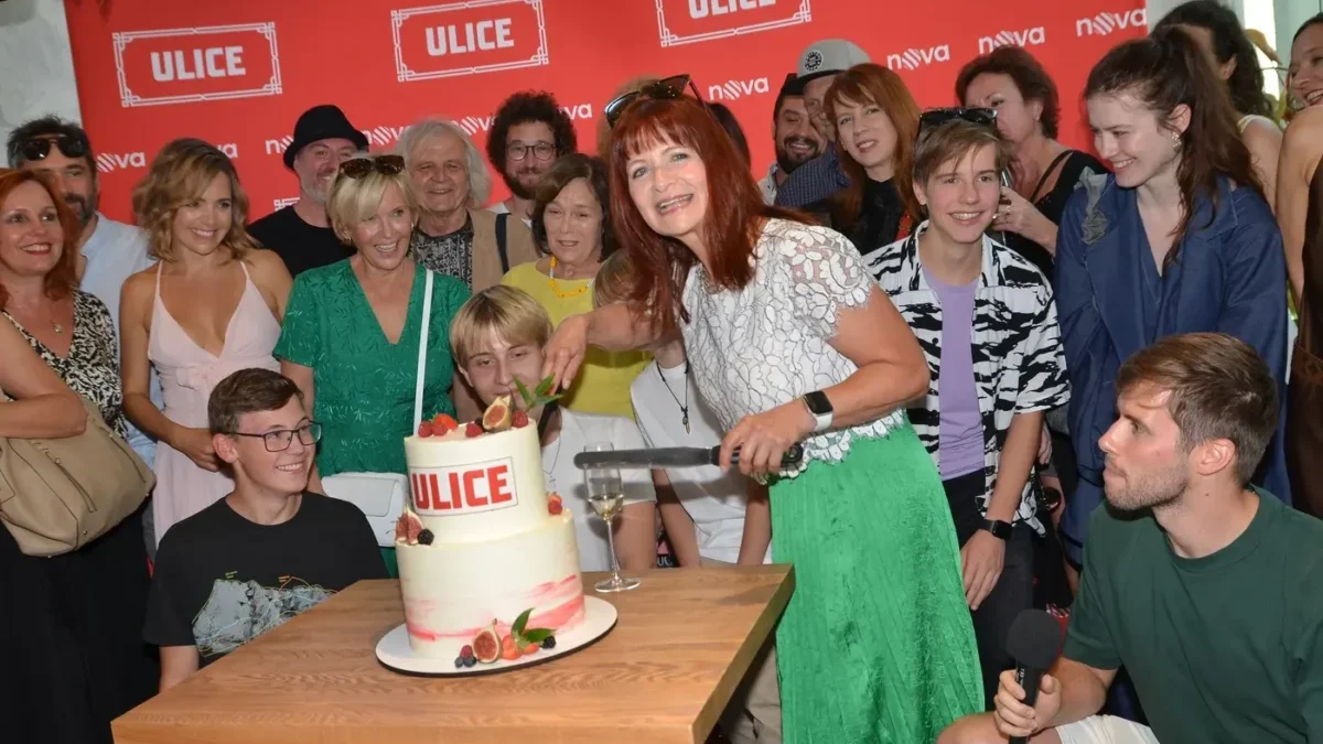 People gather around a two‑tier cake labeled ULICE as a woman in a white lace top and green skirt cuts the cake, with a group smiling in the background.