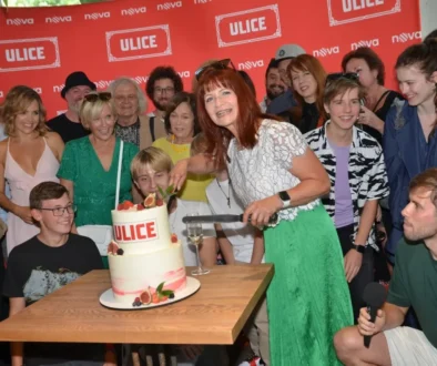 People gather around a two‑tier cake labeled ULICE as a woman in a white lace top and green skirt cuts the cake, with a group smiling in the background.