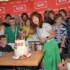 People gather around a two‑tier cake labeled ULICE as a woman in a white lace top and green skirt cuts the cake, with a group smiling in the background.