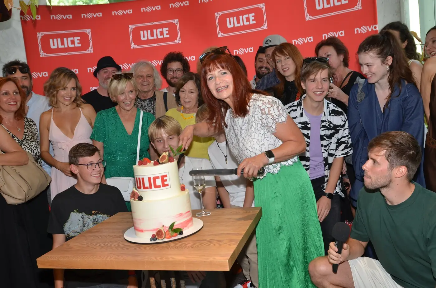 People gather around a two‑tier cake labeled ULICE as a woman in a white lace top and green skirt cuts the cake, with a group smiling in the background.