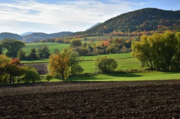 Wide rural landscape with a hillside covered in autumn trees, green fields in the valley, and a plowed field in the foreground.