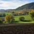 Wide rural landscape with a hillside covered in autumn trees, green fields in the valley, and a plowed field in the foreground.