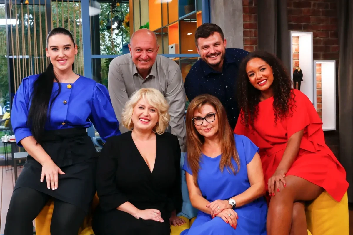 Six smiling adults posing together on a colorful TV studio set, sitting and standing in bright clothing.