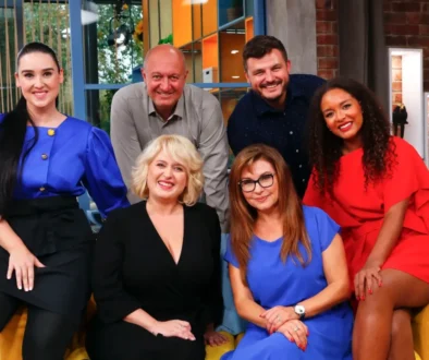 Six smiling adults posing together on a colorful TV studio set, sitting and standing in bright clothing.