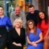 Six smiling adults posing together on a colorful TV studio set, sitting and standing in bright clothing.